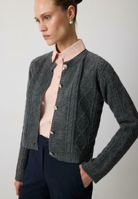 Cardigan texturat din lână gri, cu guler rotund, design cu romburi și cinci nasturi decorativi. Purtați peste o cămașă de culoare roz deschis.