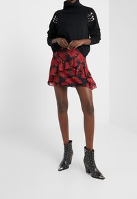 Schwarzer Rollkragenpullover mit metallischen Ösen an den Schultern, kombiniert mit einem roten, blumigen, rüschengeschmückten Minirock und schwarzen, mit Nieten verzierten Stiefeletten.