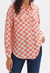 Long-sleeve shirt in cream with a coral wave pattern, featuring two chest pockets and a button-front design, styled with blue jeans.