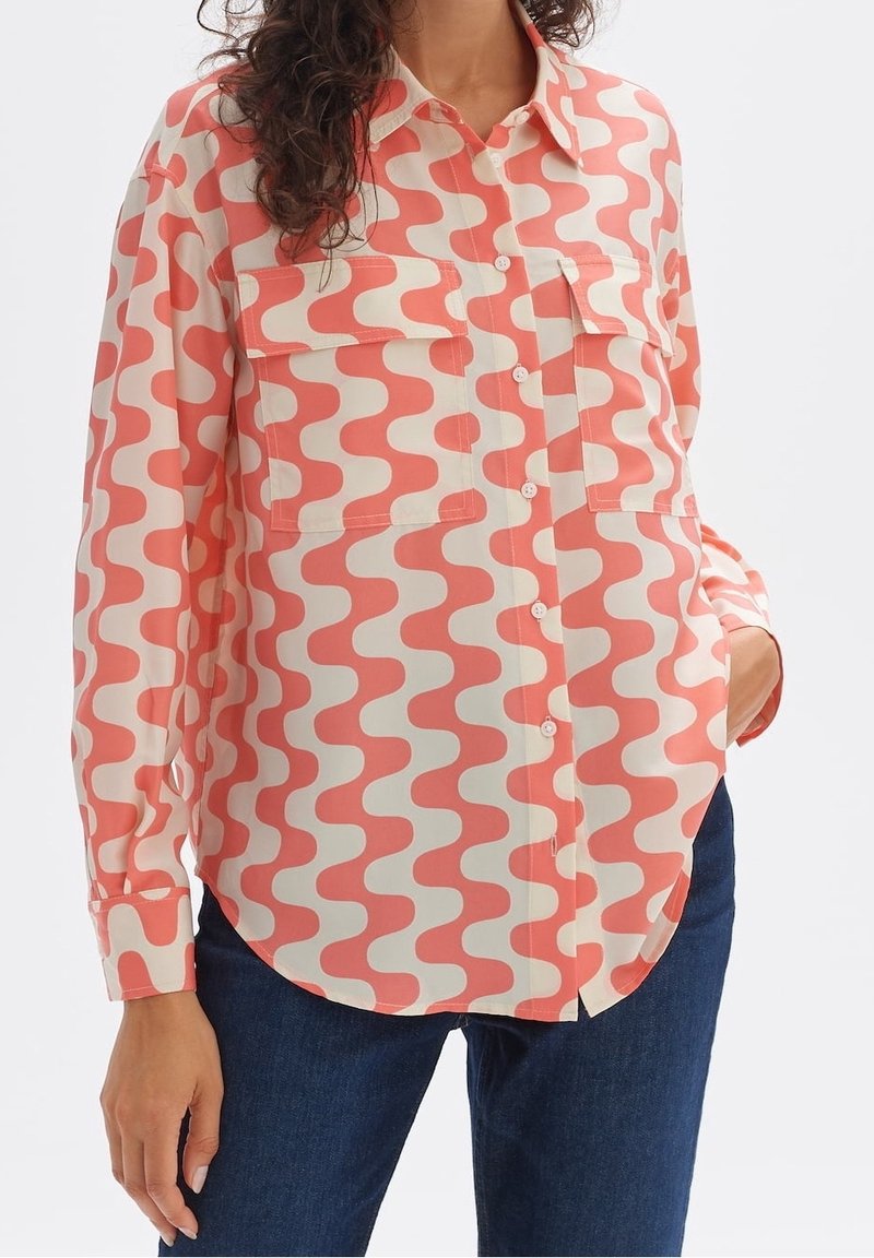 Long-sleeve shirt in cream with a coral wave pattern, featuring two chest pockets and a button-front design, styled with blue jeans.
