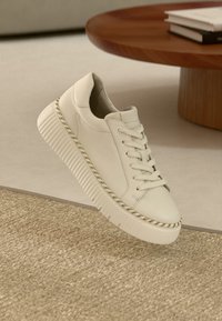 Weißer Ledersneaker mit dicker, strukturierter Sohle, rundem Zehenbereich und geflochtenem Akzent entlang des Sohlenrands. Schwebt über einer strukturierten Oberfläche.