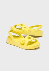 Des sandales en caoutchouc jaunes au design ouvert, avec des sangles croisées et des découpes circulaires. La semelle texturée offre adhérence et confort.