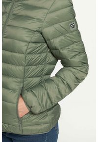 Giacca puffy verde oliva con texture trapuntata, zip frontale e tasca laterale. Presenta polsini elasticizzati e un piccolo logo sulla spalla.