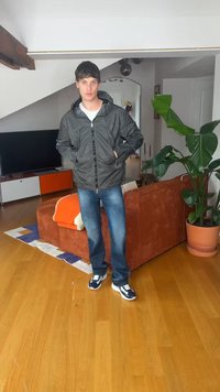 Fiatal férfi fekete dzsekiben, kék farmerral és sportcipőben egy nappaliban áll, ahol barna kanapé, televízió és nagy cserepes növény található.