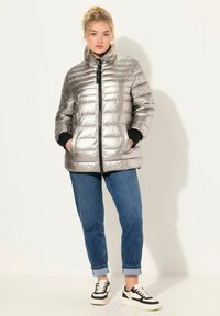 Silver puffjacka med hög krage, dragkedja fram och svarta ärmslut, kombinerad med blå jeans och svart-vita sneakers.