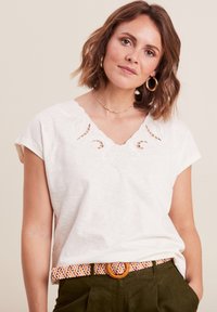 Weißes Kurzarmoberteil mit V-Ausschnitt und kunstvoller Stickerei. Kombiniert mit einem mehrfarbigen geflochtenen Gürtel und olivgrünen Hosen.