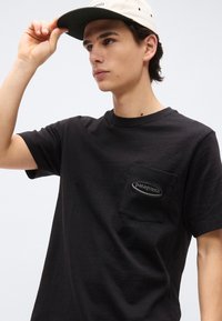 Νεαρός άνδρας που διορθώνει ένα μπεζ καπέλο, φορώντας μαύρο t-shirt με τσέπη Patagonia, ουδέτερο φόντο, συγκεντρωμένη έκφραση, κοντά σκούρα μαλλιά.