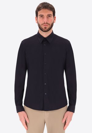 Uomo con la barba che indossa una camicia nera a maniche lunghe abbottonata e pantaloni beige, in piedi davanti a uno sfondo chiaro e uniforme.