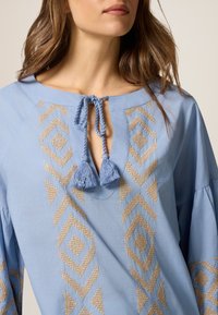 Blusa azzurra chiara con ricami geometrici dorati, scollatura con laccetto e nappine, e maniche a campana a tre quarti.