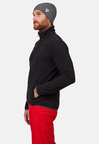 Schwarze Fleece-Trainingsjacke mit hohem Kragen, seitlichen Taschen und einem Logopatch. Graue Strickmütze mit einem dreieckigen Logodetail. Strahlend rote Hose.