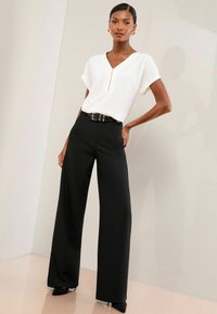 White short-sleeve top with a V-neck and gold zipper, paired with high-waisted black wide-leg trousers, accented by a black belt with a gold buckle.