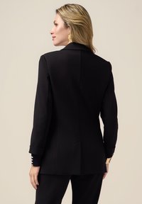 Svart blazer med en strukturerad design, slät textur, skräddarsydd passform och knappdetaljer på ärmarna, sedd bakifrån.