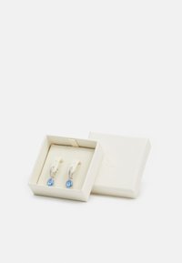 Modré kamenné náušnice v striebornom ráme, vystavené v textúrovanej krémovej krabičke s bielym interiérom. Májú jemné detaily okolo kameňov.