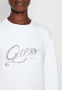 Felpa bianca con la parola "Guess" in strass argentati, con l'anno "1981" in un font semplice sottostante. Vestibilità rilassata e colletto a costine.