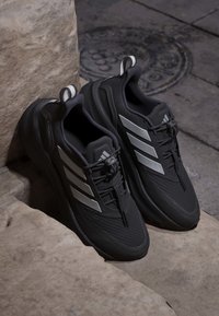 Sorte træningssko med en struktureret overflade, reflekterende sølvdetaljer og et elegant design. Inkluderer snøresystem og trækkebånd.