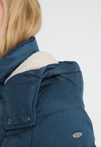 Teal pufferjakke med en sherpa-fôrvet hætte, metalliske accenter, quiltet tekstur og trykknaplukninger. Har et logo-tag på ærmet.