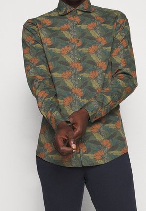 Person trägt ein langärmeliges Hemd mit grün-orangefarbenem Blumenmuster, richtet eine Manschette, kombiniert mit dunkler Hose.