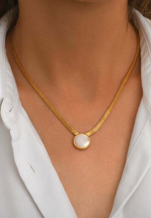 Collier chaîne en or avec un pendentif rond blanc porté autour du cou d'une personne sur une chemise blanche à col.
