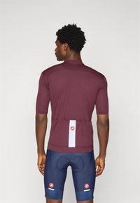 Maillot de cyclisme bordeaux à manches courtes, avec poches arrière, bande réfléchissante et détail de logo. Porté avec un short rembourré bleu. Texture lisse.