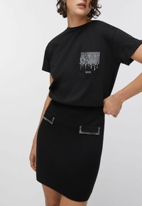 Črna majica s kratkimi rokavi, z žepom, okrašenim s kovinskimi zatiči, v kombinaciji s črno mini krilo s dvema okrasnima žepoma z zatiči.