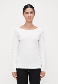 Woman with dark hair tied back wearing a plain white long-sleeve shirt and black pants, standing against a white background.