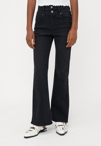 Korkeavyötäröiset mustat levenevät farkut, joissa on kaksinappinen sulku, viisi taskua ja sileä, teksturoitu pinta. Yhdistetty valkoisiin, soljellisiiin kenkiin.