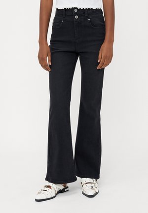 Højtaljede sorte flagrende jeans med en to-knaps lukning, fem lommer og en glat, tekstureret overflade. Bæres med hvide, spænde sko.