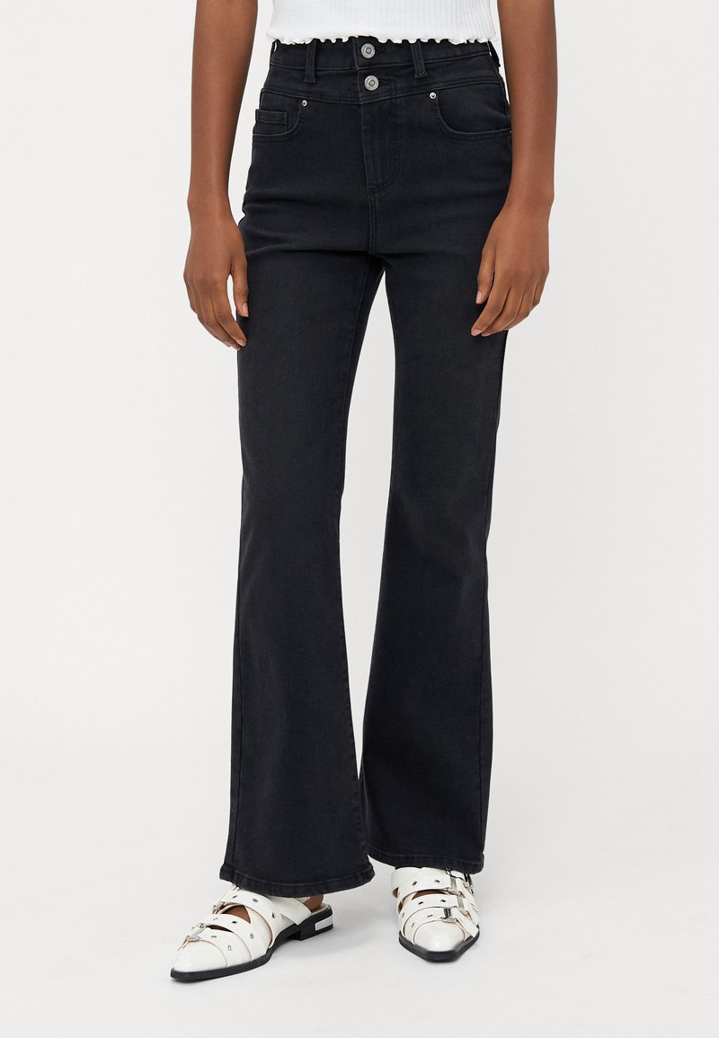 Korkeavyötäröiset mustat levenevät farkut, joissa on kaksinappinen sulku, viisi taskua ja sileä, teksturoitu pinta. Yhdistetty valkoisiin, soljellisiiin kenkiin.