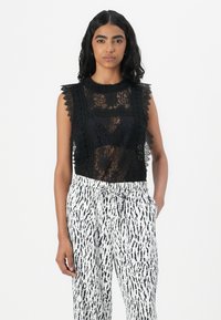 Top senza maniche in pizzo nero con scollatura a scallop e motivi intricati, abbinato a pantaloni bianchi con stampa astratta nera.