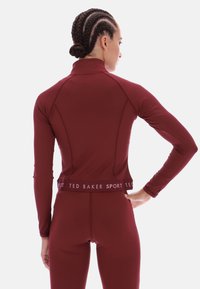 Vīna sarkanā sporta krekls ar augstu apkakli, gariem piedurknēm un piegulošu dizainu, ar zīmola jostu un gludu tekstūru.