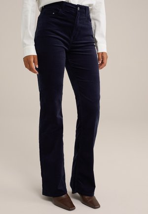 Navy fluwelen uitlopende broek met een hoge taille, voorzien van voorzakken en een subtiele textuur. Gdragend met een wit shirt met lange mouwen en bruine schoenen.