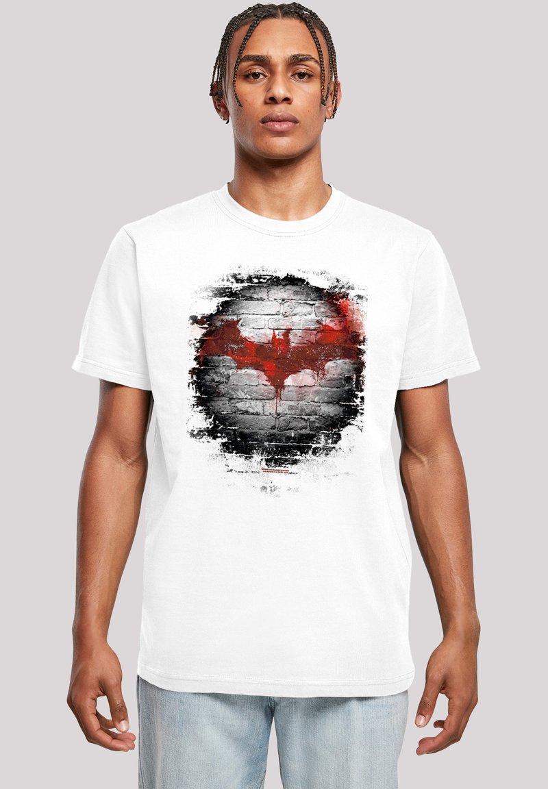 Hvid bomulds t-shirt med et slidt rødt og sort flagermus-symbol på et murstensvæg-grafik. Korte ærmer, rund hals, afslappet pasform.