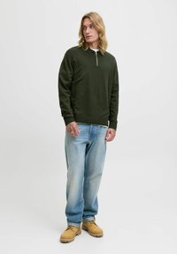 Mørkegrøn, lynlås-pullover med krave; båret med lyseblå, løstsiddende jeans og brune arbejdssko. Glat tekstur, afslappet pasform.