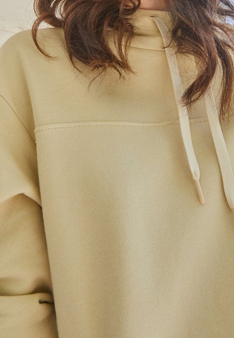 Beige hoodie tillverkad av mjukt tyg. Har en central söm och dragsko med plastspetsar. Enkelt, avslappnat design.
