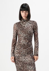 Woman with dark straight hair wearing a long-sleeve, leopard-print fitted dress and a pearl necklace, standing against a white background.