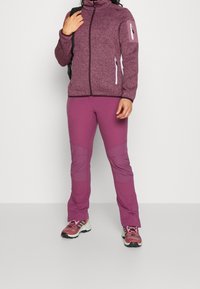 Violetti fleece-takki vetoketjullisten taskujen kanssa, yhdistetty violetteihin housuihin, joissa on polviakselit. Yhdistetty monivärisiin urheilukenkiin.