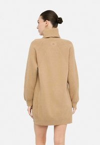 Abito maglione beige con colletto alto, texture a coste, maniche lunghe e dettaglio del logo sul retro. Vestibilità normale con spacco laterale.