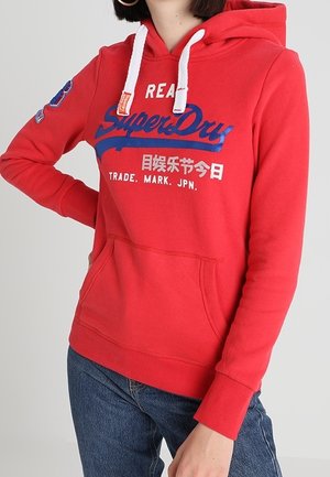 Czerwona bluza z kapturem z białymi sznurkami, niebiesko-białe logo SuperDry i japońskie znaki, kieszeń z przodu, noszona z niebieskimi dżinsami.
