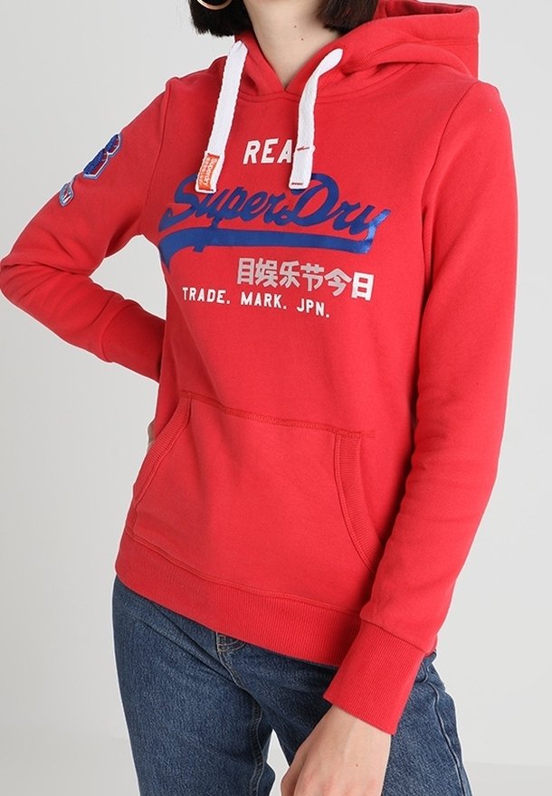 Sweat à capuche rouge avec cordons blancs, logo SuperDry bleu et blanc et caractères japonais, poche kangourou à l'avant, porté avec un jean bleu.
