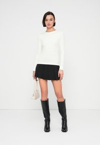 Λευκό τοπ μακρυμάνικο, μαύρη πλισέ μίνι φούστα, μαύρες μπότες μέχρι το γόνατο και μικρή λευκή τσάντα, τοποθετημένα σε απλό φόντο.