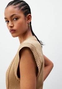 Beežist kootud ilma varrukateta topp, ribakujundusega, laiad õlad ja ümmargune kaelus. Juuksed on punutud, minimaalne ehtelaad.