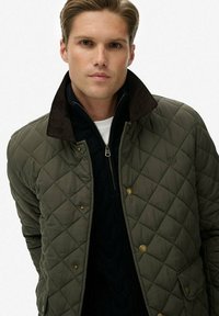 Giacca trapuntata verde oliva con colletto in corduroy e bottoni dorati, abbinata a un maglione navy sottostante per calore e texture.