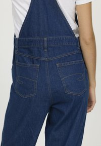 Salopette en denim bleu foncé avec des coutures visibles et deux poches arrière comportant des détails de couture décoratifs. Coupe décontractée et bretelles ajustables.