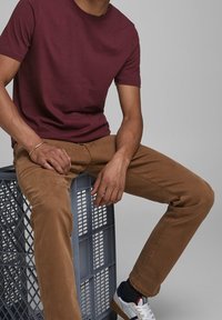 Un homme portant un t-shirt bordeaux, un pantalon marron et des baskets blanches assis sur une caisse en plastique grise contre un fond uni.