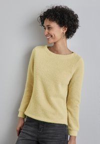 Maglione giallo testurizzato con scollo a girocollo e maniche lunghe, abbinato a jeans aderenti grigio scuro. Caratterizzato da una finitura morbida e pelosa.