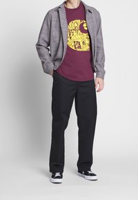 Maroonowa koszulka graficzna z żółtym nadrukiem, połączona z czarno-białą koszulą w kratę, czarnymi spodniami i czarnymi sneakersami z białymi akcentami.