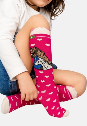 Kind sitzt mit überkreuzten Beinen und trägt leuchtend pinke Socken, die mit weißen Herzen und einem Motiv eines Mädchens mit einem Hund verziert sind.