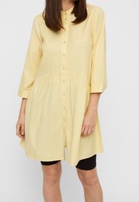 Gelbes Tunika-Kleid mit Knopfleiste, ausgestelltem Saum und Dreiviertelärmeln, aus leichtem Stoff und mit lockerem Schnitt.