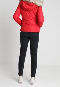 Rød polstret jakke med en hætte foret med pels, båret over sorte jeans og hvide sneakers. Stoffet ser glat ud med en blank finish.