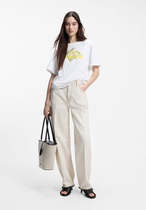 Femme aux longs cheveux foncés portant un t-shirt blanc à motif, un pantalon large beige, des sandales à talons noires et tenant un sac fourre-tout beige et noir.